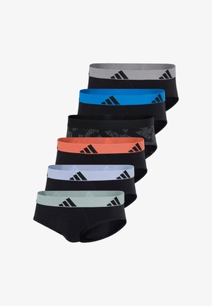 Athletischer Mann steht barfuß und trägt schwarze Unterhosen mit orangefarbenem Adidas-Bund vor einem schlichten weißen Hintergrund.