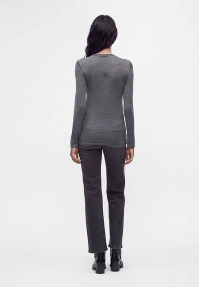 Femme aux longs cheveux foncés portant une chemise grise cintrée à manches longues, un jean noir et des bottes à talons noires, debout de dos.