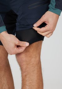 Marineblaue Shorts mit einer Netzunterlage, einer Akzenttasche und einem schlanken Design; Handanpassung des unteren Saums, der schwarzes Netzmaterial freilegt.