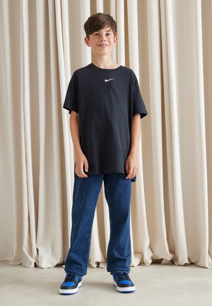 T-shirt noir à manches courtes avec un logo Nike blanc, associé à un jean en denim bleu et des baskets noires avec des accents bleus, présentées devant des rideaux neutres.