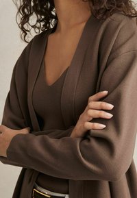Cardigan marrone a maglia con maniche lunghe, apertura frontale e tessuto texturizzato. Indossato sopra un top a V coordinato, mostrando una palette di colori coerente.
