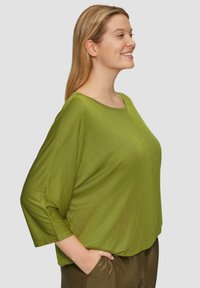 Jonge vrouw die glimlacht, met een losse groene trui en een bruine broek, met één hand in de zak, tegen een effen achtergrond.