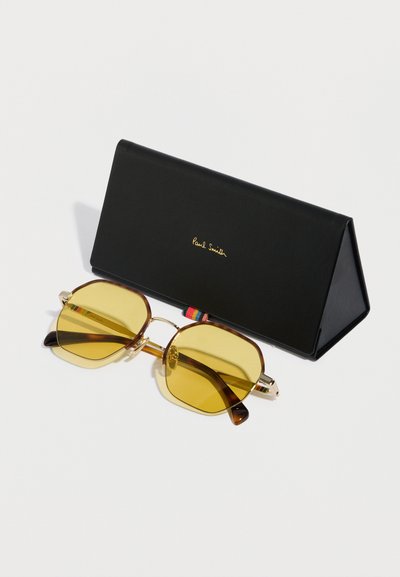 Occhiali da sole con lenti gialle, dettagli in oro e montatura in tartaruga marrone, posizionati accanto a una custodia triangolare nera con scritte dorate.