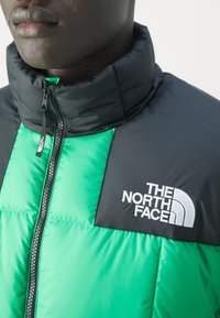 Zelená a černá péřová bunda s vysokým límcem, zipovým zapínáním a logem "The North Face" vyšívaným bílou nití na hrudi.