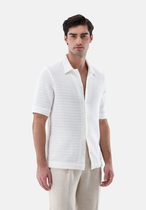 Junger Mann trägt ein weißes, kurzärmeliges, strukturiertes Hemd mit offenem Kragen und hellbeige Hose, steht vor einem schlichten, hellen Hintergrund.