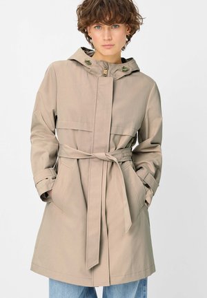 Frau mit kurzen lockigen Haaren, die einen beigen, taillierten Kapuzenmantel mit Taschen trägt und vor einem schlichten weißen Hintergrund steht.