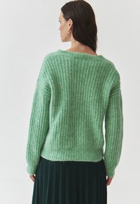 Pull en maille côtelée vert, avec une coupe décontractée et un col rond, présentant une texture douce et duveteuse. Porté sur une jupe plissée noire.
