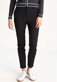 Pantalons noirs sur mesure avec une coupe slim, en tissu lisse, présentant un pli à l'avant et des poches latérales, associés à des baskets blanches.