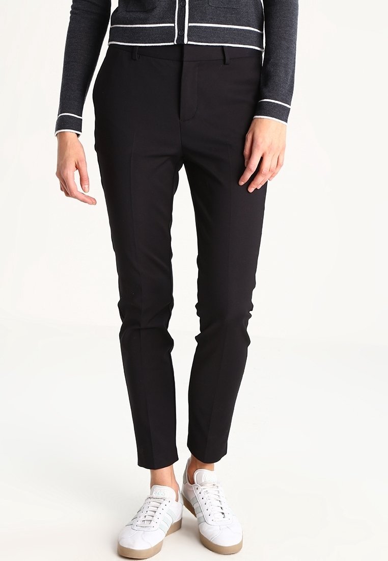 Pantalons noirs sur mesure avec une coupe slim, en tissu lisse, présentant un pli à l'avant et des poches latérales, associés à des baskets blanches.