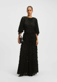 Abito nero con un design testurizzato e stratificato. Maniche lunghe, vestibilità ampia e vita affusolata. La modella tiene una piccola borsa nera.