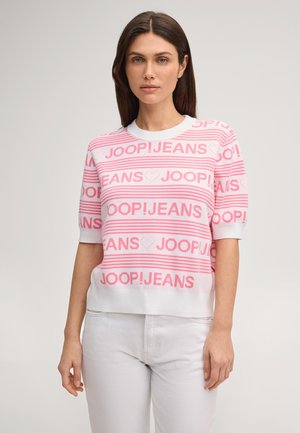 Femme portant un pull à manches courtes rayé blanc et rose avec le texte "JOOP! JEANS" et des motifs de cœurs, assorti à un pantalon blanc.
