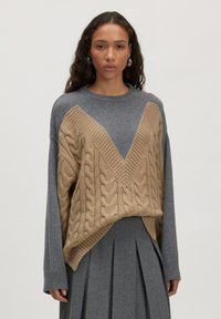 Vrouw in een grijze gebreide trui met een beige kabelgebreide V-vormige overlay en een bijpassende grijze plissérok, staande tegen een effen achtergrond.
