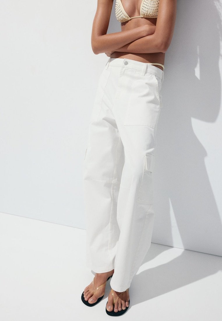Femme debout portant un pantalon cargo blanc, un haut de bikini beige clouté et des tongs noires avec des brides transparentes sur fond blanc.