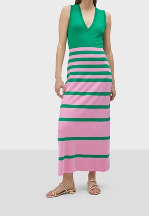 Femme portant un haut vert sans manches et une jupe maxi rose avec des rayures horizontales vertes, debout avec des sandales dorées sur fond blanc.