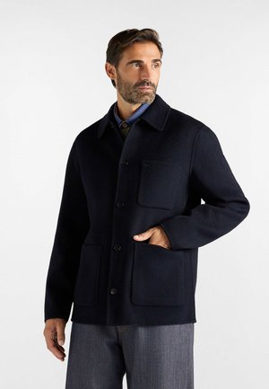 Uomo che indossa una giacca di lana blu scuro con bottoni e tre tasche frontali, in piedi con una mano in tasca, guardando di lato.