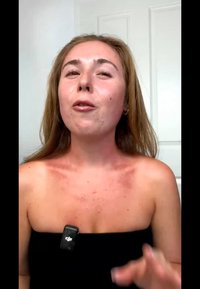 Haut noir sans bretelles, peau claire avec une légère rougeur aux épaules, parlant face à la caméra, portant un micro-cravate noir sur la poitrine.