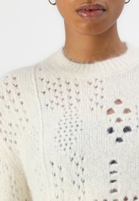 Vit stickad tröja med ett texturerat mönster med synliga hål, accentuerad av ljusbruna och svarta prickar genom hela designen.