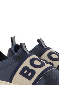 Chaussures bateau navy à enfiler avec des sangles élastiques beiges, arborant des lettres navy audacieuses. Tissu texturé avec une finition lisse et peu de quincaillerie visible.