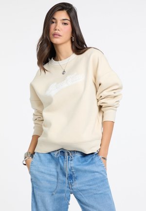 Sweatshirt beige avec un col rond et des poignets côtelés, orné d'un logo brodé blanc. Associé à un pantalon en denim bleu à coupe décontractée.