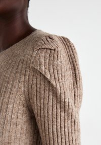 Maglione a coste beige con design aderente e dettaglio arricciato sulle spalle, che presenta una texture morbida e sottili variazioni di colore.