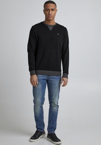 Schwarzer Pullover mit langen Ärmeln, V-Ausschnitt und gerippten Bündchen, getragen über einem grauen Rundhalsausschnitt-Shirt, kombiniert mit hellblauen Jeans und schwarzen Sneakern.