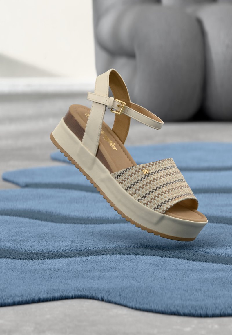 Sandale à plateforme beige avec une bride avant tissée et une boucle cheville, présentée au-dessus d'une surface bleue texturée avec une sculpture grise floue en arrière-plan.