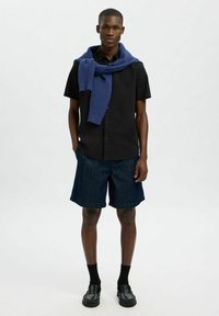 Camisa negra de manga corta, suéter azul de punto drapeado sobre los hombros, pantalones cortos de mezclilla oscura, zapatos negros y calcetines negros. Diseño simple y limpio.
