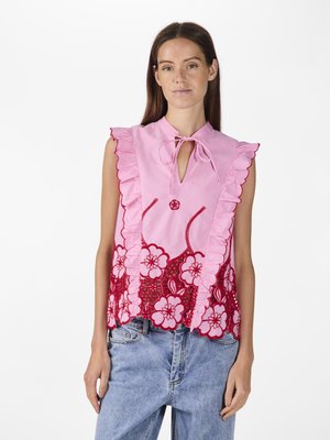 Pink blouse with ruffled edges, floral lace detailing in red, round button embellishment, and tie neck closure. Worn with denim jeans.