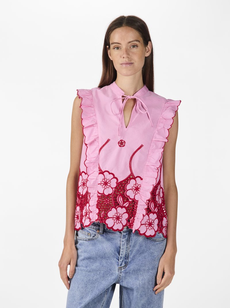 Blusa rosa con bordi arricciati, dettagli in pizzo floreale rosso, decorazione con bottone tondo e chiusura con nodo. Indossata con jeans di denim.