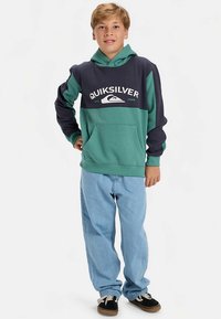 Zöld és tengerészkék kapucnis pulóver kenguruzsebbel és fehér logós felirattal. Világoskék farmerrel és fekete cipővel párosítva.