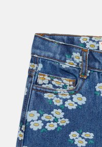 Μπλε τζιν ύφασμα με λευκά και κίτρινα λουλούδια μαργαρίτας, με ζώνη μέσης, θηλιές ζώνης και λεπτομέρειες εμπρόσθιας τσέπης με πορτοκαλί ραφές.