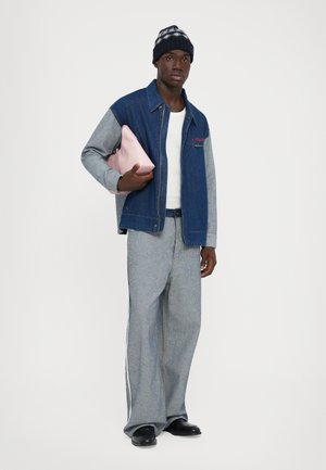 Un homme portant une veste bleue et grise, un pantalon large gris, des chaussures noires, un bonnet en tricot, tenant une pochette rose contre un fond uni.