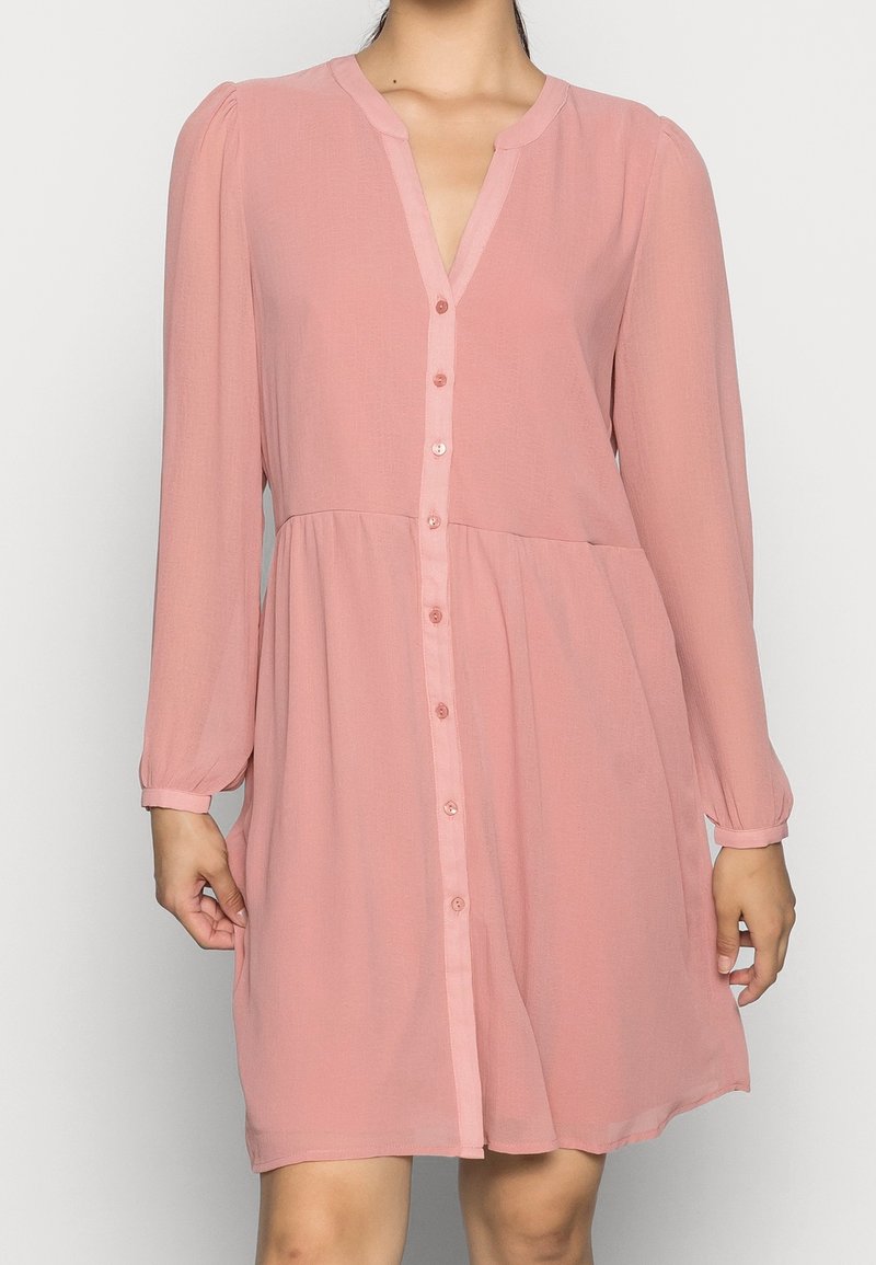 Femme portant une robe rose ample à manches longues, boutonnée, avec un col en V et une taille froncée.