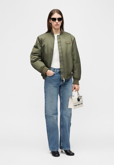 Άτομο που φοράει μαύρα γυαλιά ηλίου, χακί bomber μπουφάν, λευκό τοπ, μπλε τζιν, μαύρα παπούτσια, κρατώντας μικρή λευκή τσάντα χειρός με κείμενο.