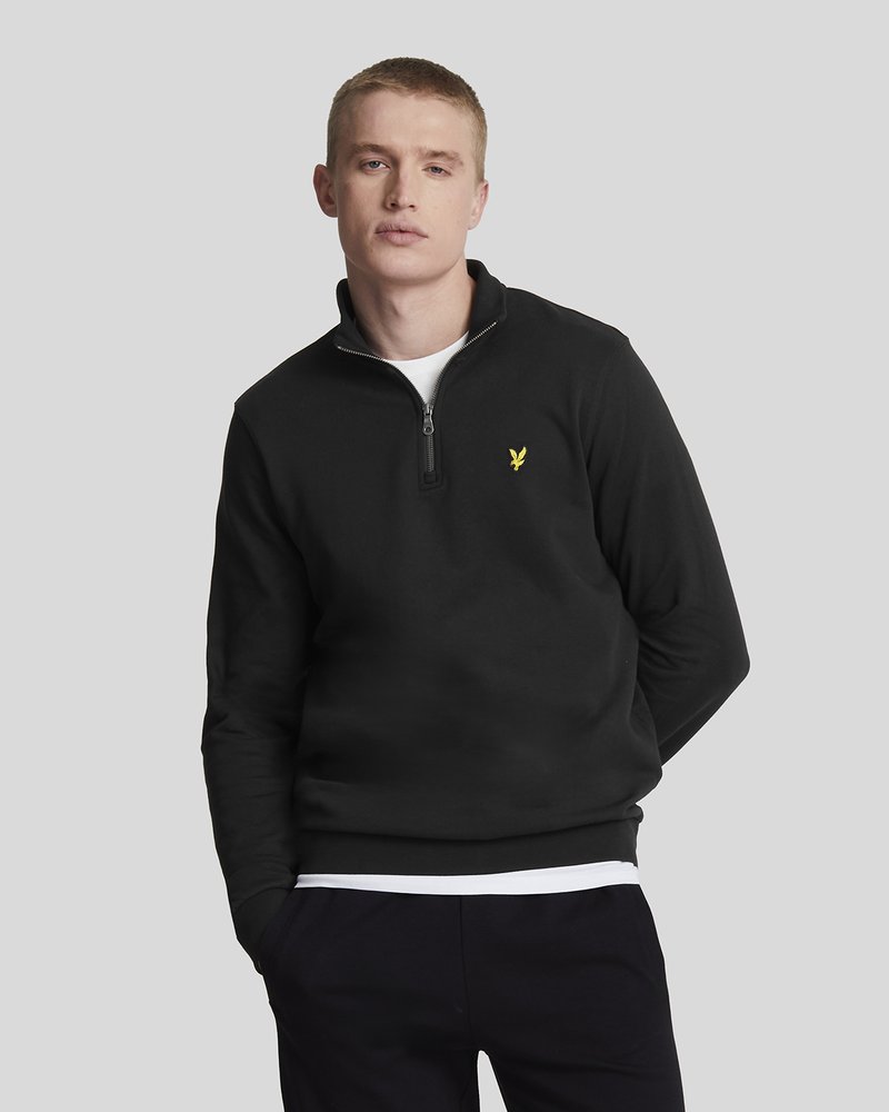 Jonge man in een zwart sweatshirt met rits en een klein geel vogellogo, witte onderhemd en zwarte broek voor een lichtgrijze achtergrond.