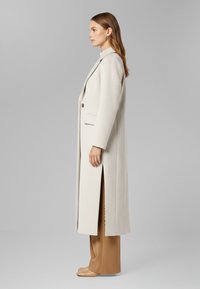 Cappotto lungo, beige chiaro, realizzato in tessuto testurizzato, con un design tailleur, chiusura a un solo bottone e spacchi laterali. Indossato sopra pantaloni a gamba larga marroni.