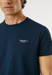 Muž nosiaci tmavomodré tričko Pepe Jeans London, viditeľný od krku po polovicu trupu, na jednoduchom svetlom pozadí.