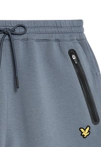Pantaloni grigi in felpa con vita elasticizzata, cordino e tasca nera con zip. Presenta un piccolo logo giallo di un uccello. Texture liscia.
