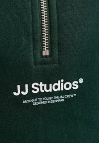 Tissu vert foncé avec une fermeture éclair argentée et un texte blanc indiquant "JJ Studios, présenté par l'équipe J&J, conçu au Danemark."