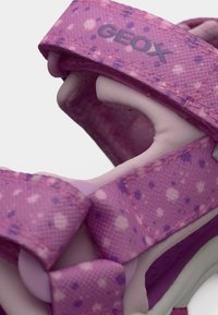Gros plan sur une sandale pour enfants rose et violette avec des brides réglables et le nom de la marque Geox sur la bride supérieure.