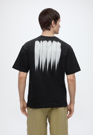 Homme de dos portant un t-shirt noir avec un texte stylisé blanc dans le dos et un pantalon kaki sur un fond uni clair.