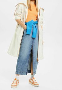 Světle krémový trenčkot, oranžový žebrovaný top, džíny s vysokým rozparkem, modrý svetr svázaný v pase a tenisky s oranžovými akcenty.