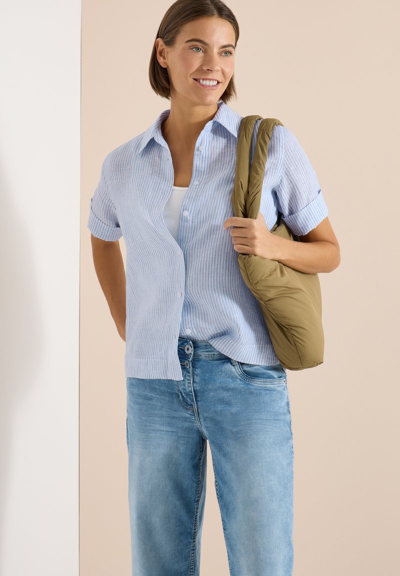 Jeune femme portant une chemise à rayures bleu clair et un jean, souriante, avec un sac à bandoulière matelassé vert olive sur l'épaule droite.