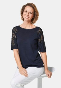 Marineblauwe trui met korte mouwen, voorzien van zwarte kantaccenten en versieringen. Soepele textuur, ronde halslijn en een ontspannen pasvorm.
