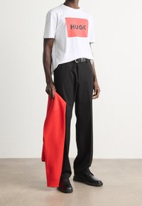 Camiseta blanca con el logotipo rectangular rojo "HUGO", pantalones negros, zapatos negros y un suéter rojo colgado de un brazo.