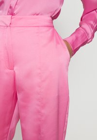 Calças de cetim cor-de-rosa com uma textura suave, ajuste feito à medida, bolsos laterais e um design de cintura alta. Detalhes de costura visíveis realçam a estrutura.