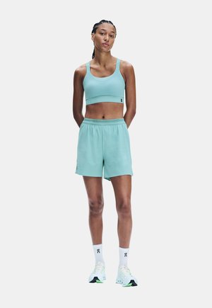 Femme athlétique debout, mains dans le dos, portant un soutien-gorge de sport bleu clair, un short assorti, des chaussettes blanches et des chaussures de course blanches.