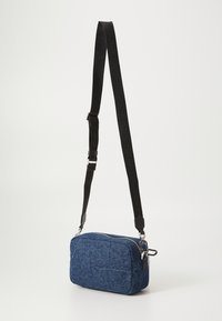 Blaues Denim-Crossbody-Tag mit einem Reißverschluss, ausgestattet mit einem verstellbaren schwarzen Gurt und silbernen Hardware-Details. Kompakte und rechteckige Form.