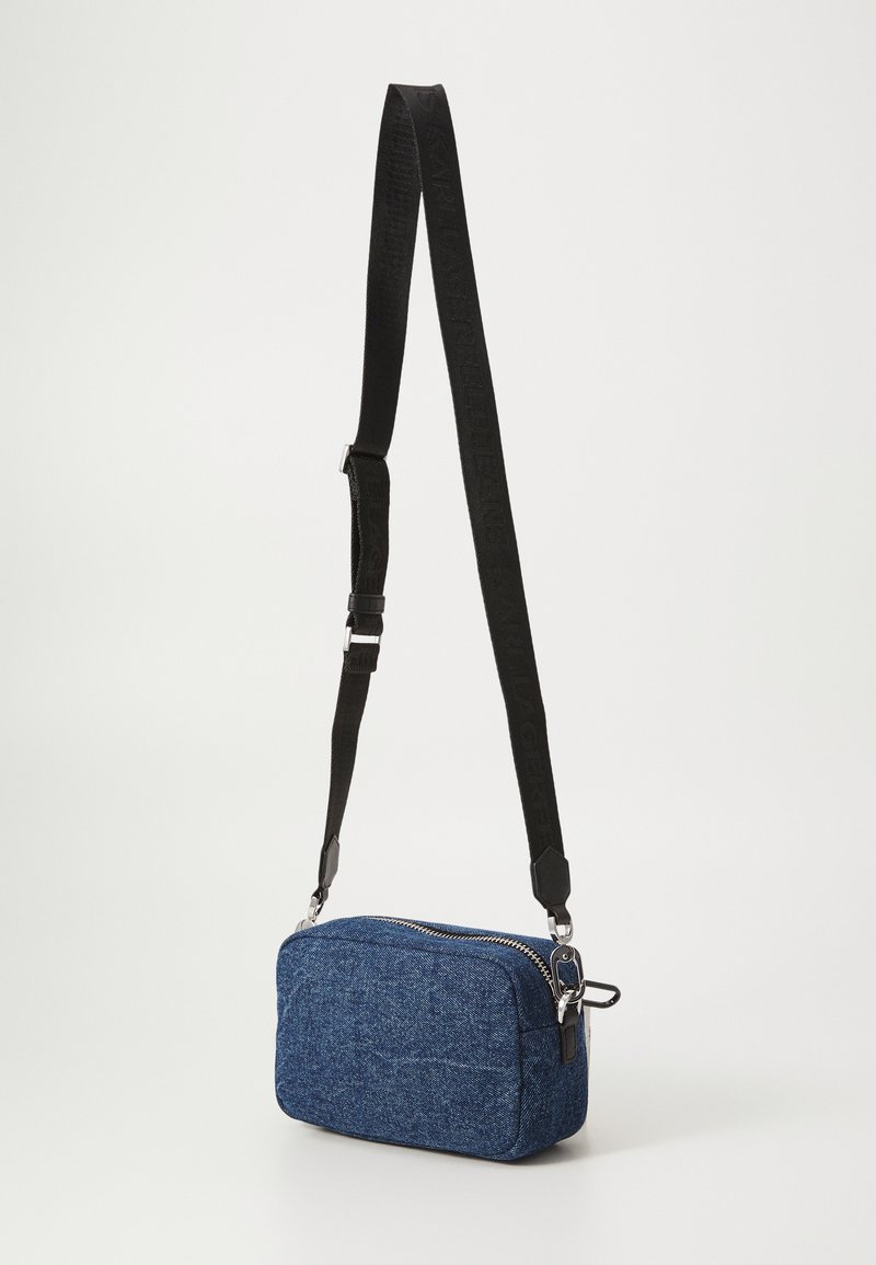 Blaues Denim-Crossbody-Tag mit einem Reißverschluss, ausgestattet mit einem verstellbaren schwarzen Gurt und silbernen Hardware-Details. Kompakte und rechteckige Form.