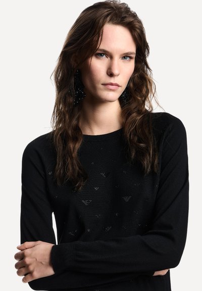 Maglione nero lavorato a maglia con motivo a rilievo e sottili accenti lucidi. Maniche lunghe e scollo rotondo, con capelli di media lunghezza.